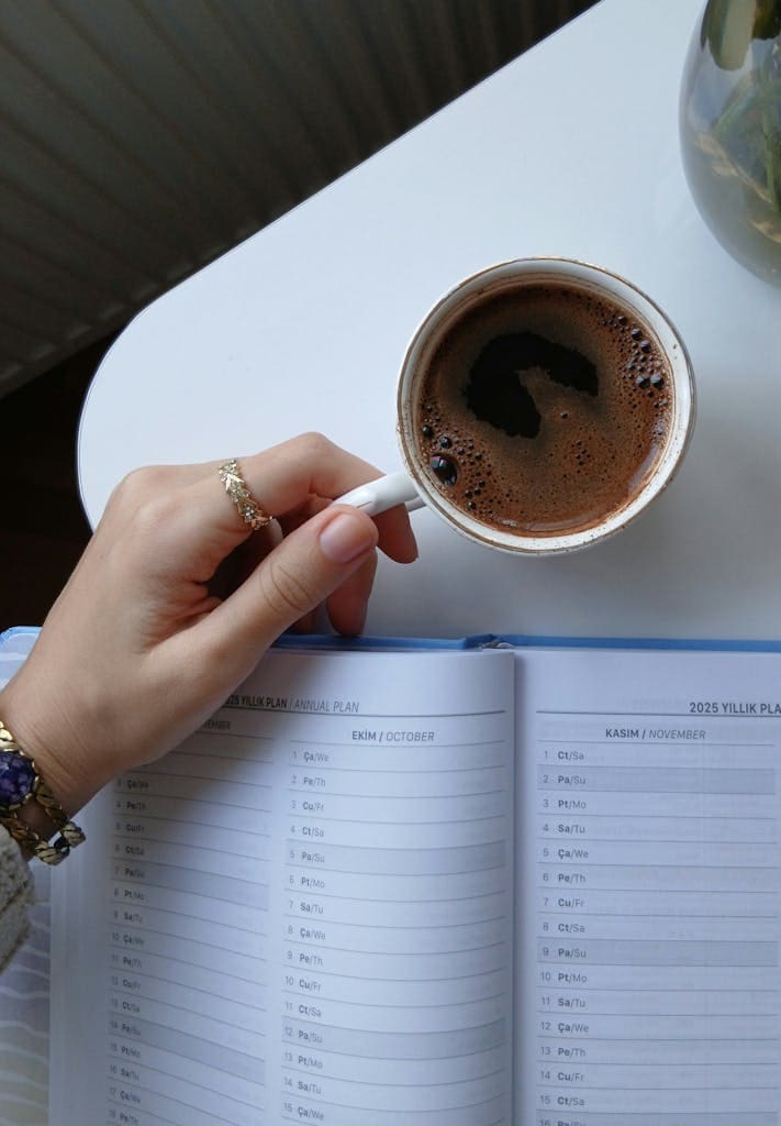 Top Tips When Searching for Cheap Flights | Imagine Greece Retreats Close-up of a hand holding a coffee cup above an open planner with visible dates.